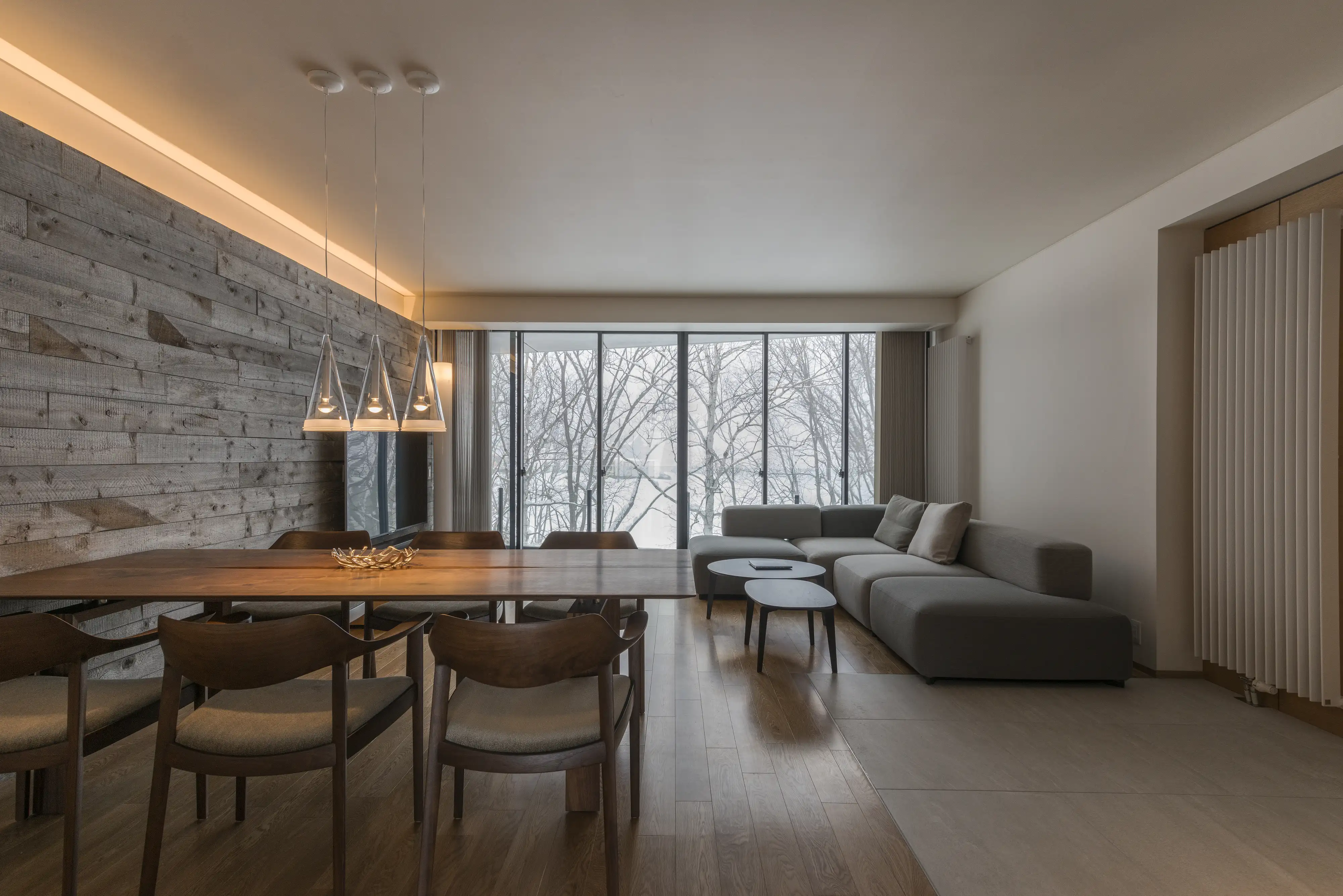 Open-plan living space with wood dining table, grey sofa, whitewashed wood accent wall, and floor-to-ceiling windows