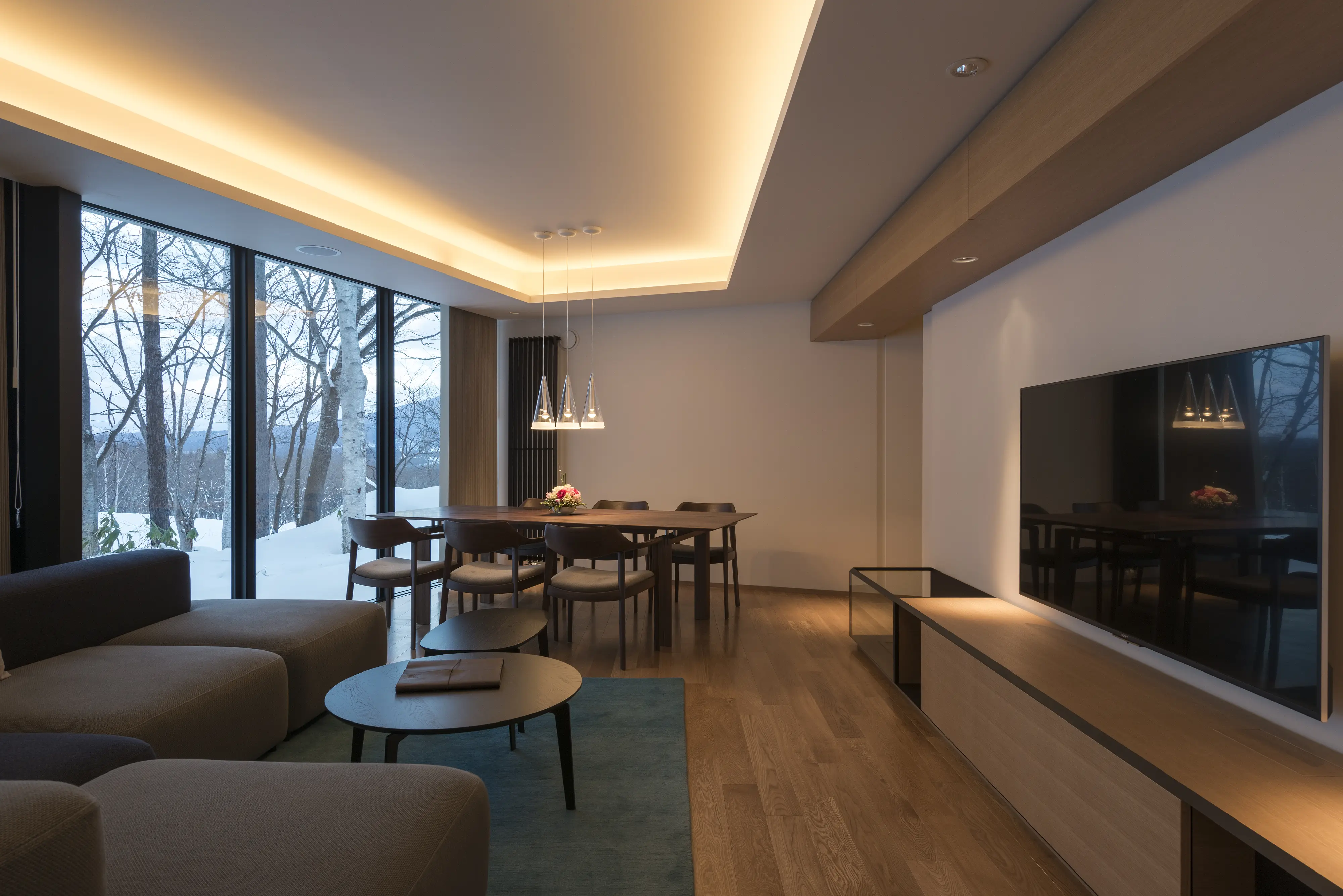 Open-plan living and dining area with floor-to-ceiling windows overlooking snowed trees, wooden dining table with chairs,