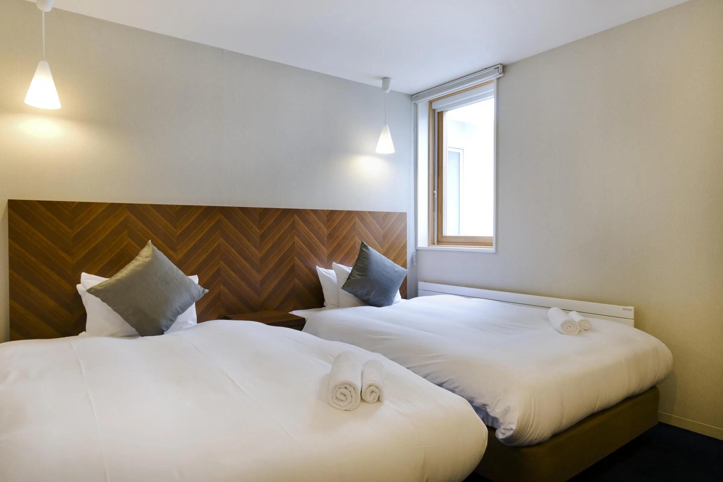 Two single beds with herringbone timber headboard, white bedding and rolled towels, pendant lights above, window with wooden